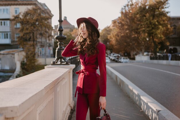 Cute beautiful stylish woman in purple suit walking in city street, spring summer autumn season fashion trend wearing hat, holding purse