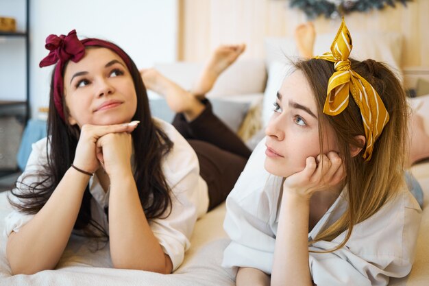 Cute beautiful teen girls wearing head bands lying on sofa, holding hands under chin and looking with thoughtful facial expressions, feeling bored