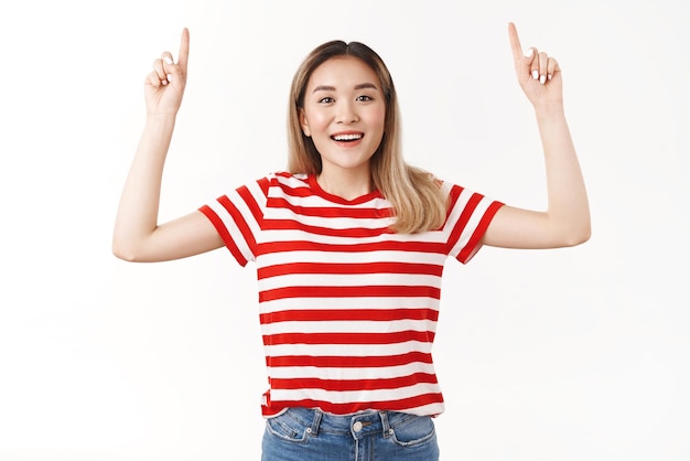 Cute blond asian girl suggest perfect vacation trip pointing up index fingers travel agency promo smiling broadly look excitement joy camera standing red striped tshirt white background