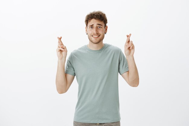 cute blond european male student in t-shirt raising crossed fingers and smiling with shy and awkward expression