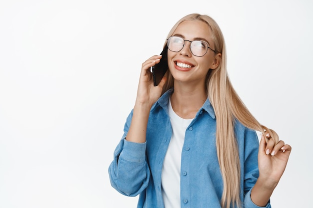 白い背景の上に立っている電話を持っている携帯電話でいちゃつくと話している眼鏡のかわいいブロンドの女の子セルラー技術の概念