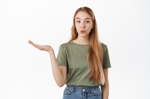 Cute blond girl holding empty hand, raising arm with opened palm as if display product, advertising at item, looking intrigued at front, standing over white wall