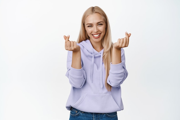 Cute blond girl in hoodie showing finger heart gesture, smiling and looking lovely, standing in purple hoodie on white