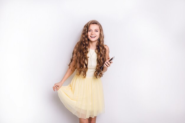 Cute blond girl listening to music on earphones on smartphone is having fun. She smiles widely and plays with her yellow dress. She had long curly blond hair