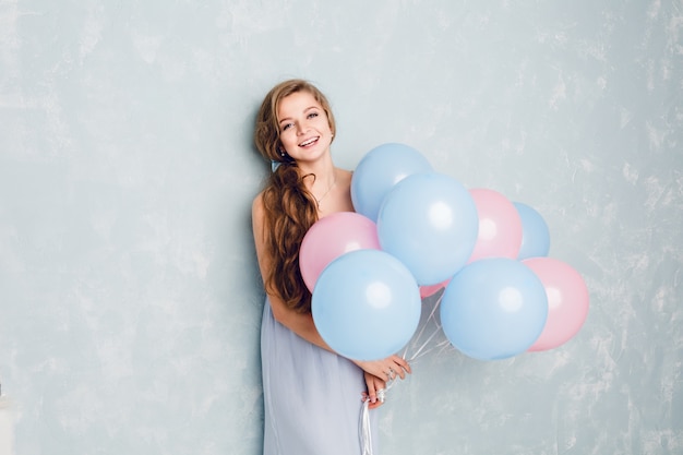 Cute blond girl standing in a studio, smiling and holding blue and pink balloons.