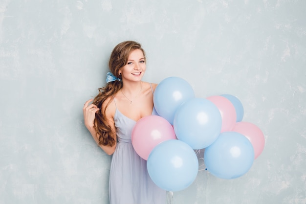 Cute blond girl standing in a studio, smiling and holding blue and pink balloons.