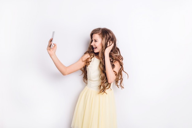 Cute blond girl taking a selfie on smartphone and having fun. She smiles widely and plays with her hair. She wears yellow dress. She had long curly blond hair