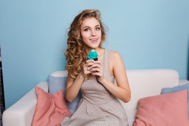 Милая белокурая девушка с вьющимися волосами, сидя на софе и держа голубой кекс.