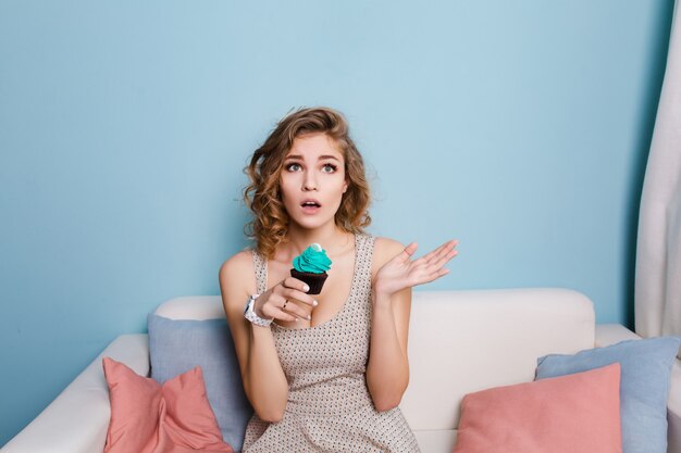 Cute blond girl with curly hair sitting on a sofa and holding a blue cupcake.