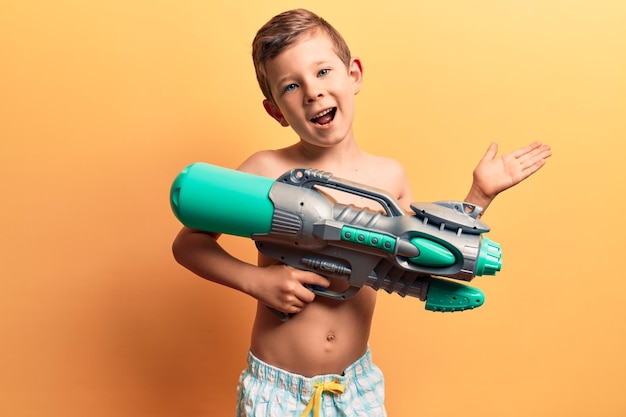 Cute blond kid wearing swimwear holding water gun celebrating achievement with happy smile and winner expression with raised hand