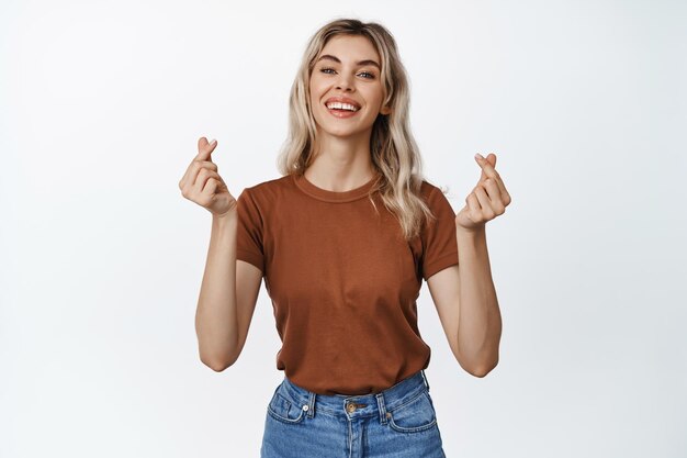 Cute blond woman smiling showing finger hearts gesture standing in casual clothes against white background