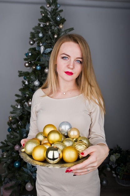 Cute blonde girl on beige dress against new year tree with with golden new year balls at studio Happy holidays