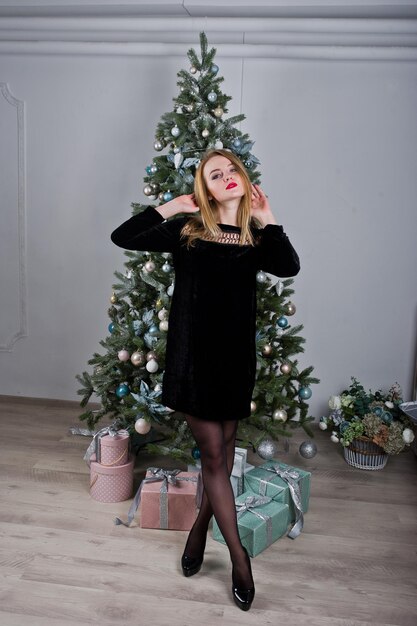 스튜디오에서 크리스마스 선물과 함께 새 해 나무에 대 한 검은 드레스에 귀여운 금발 소녀 해피 홀리데이