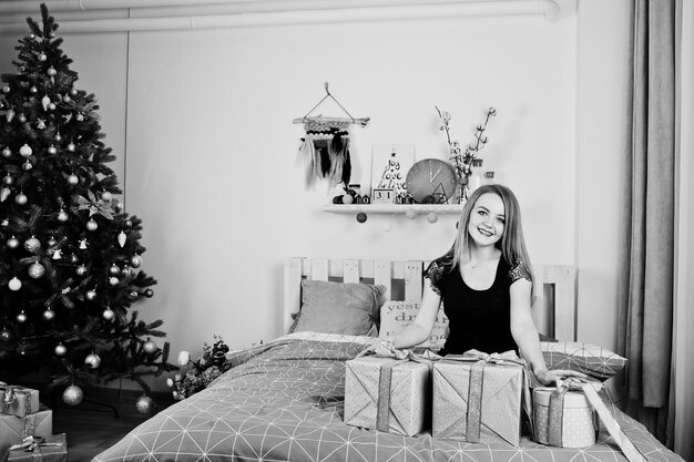 Cute blonde girl on black dress lying on bed with gifts boxes against new year tree at studio Happy holidays