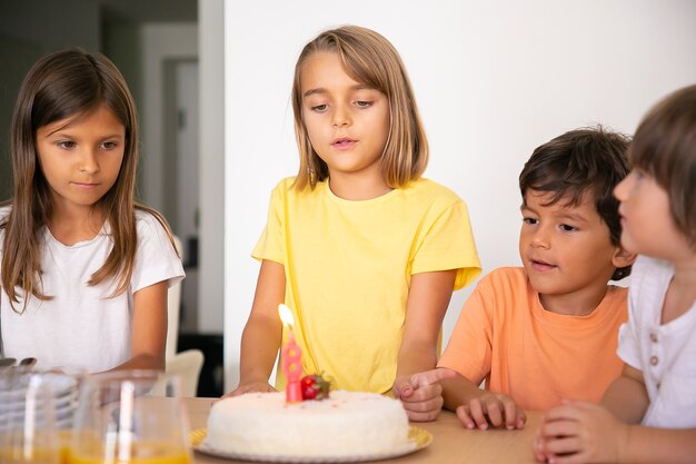 Cute blonde girl making wish and celebrating her birthday with friends. Lovely children standing in room together and looking at tasty cake with candle. Childhood, celebration and holiday concept