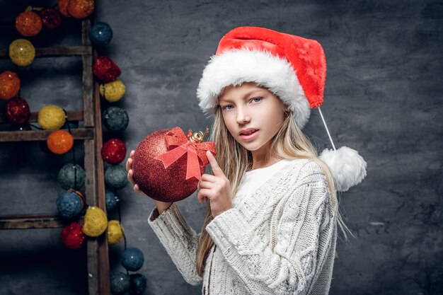 산타 모자를 쓴 귀여운 금발 소녀가 빨간 크리스마스 공을 들고 있습니다.