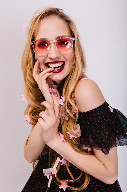 Cute blonde girl with finger in mouth, looking happy and cheerful, photoshoot at party. Has pretty curly hair, beautiful smile. Wearing stylish black dress and pink glasses. Isolated..
