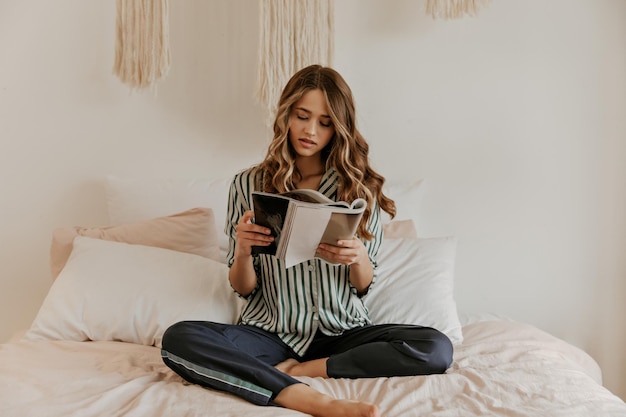 無料写真 シルクの縞模様のパジャマでかわいい金髪の長い髪の巻き毛の女性は、雑誌を読んで、快適に座っています