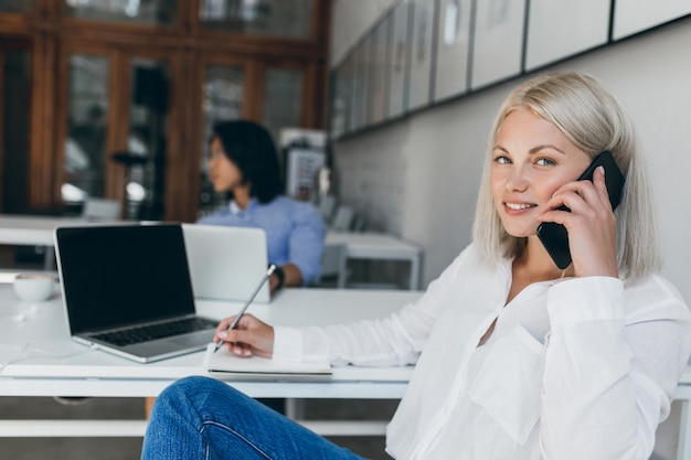 Cute blonde secretary in white blouse talking on phone and writing data in notebook. Indoor portrait of long-haired asian it-specialist with graceful lady at reception desk.