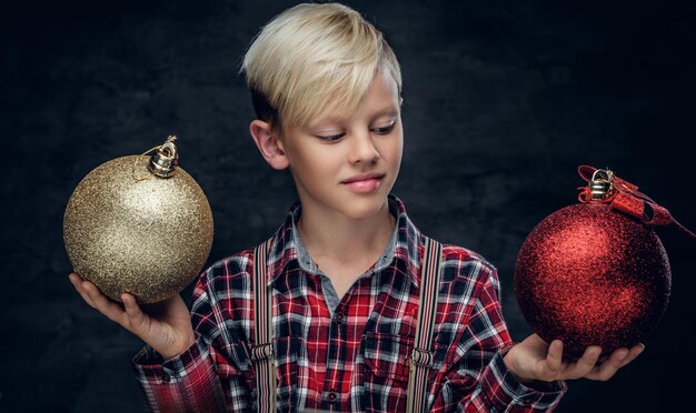 귀여운 금발 10대 소년이 금색과 빨간색 크리스마스 공을 들고 있습니다.