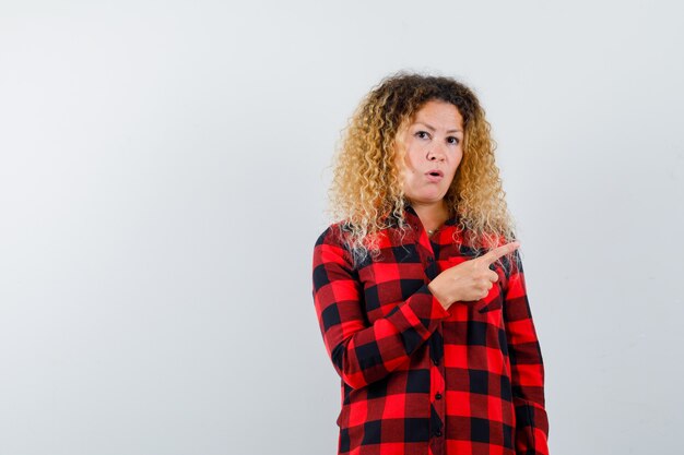 Cute blonde woman pointing right in checked shirt and looking puzzled , front view.