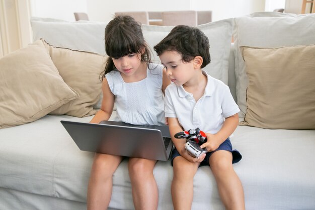 Cute boy and girl sitting on couch at home and using laptop, watching video or movie.