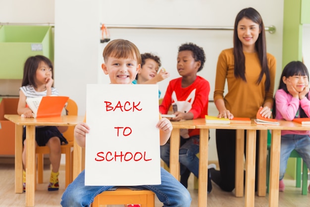 Free photo cute boy holding back to school poster with happy face in kindergarten classroom, kindergarten education concept, vintage effect style pictures.