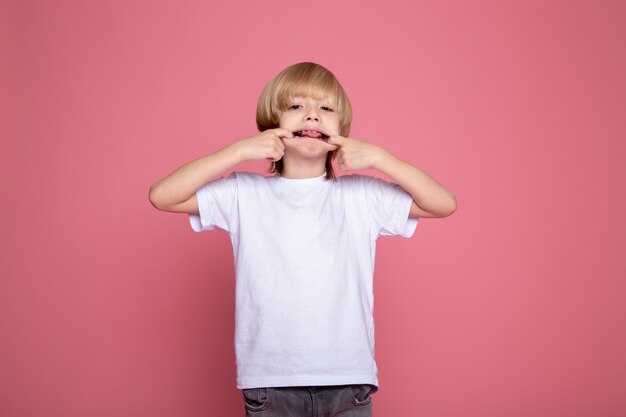 ピンクの机の上のかわいい愛らしい子少年の白いtシャツとグレージーンズの肖像画で面白い表情を作るかわいい男の子