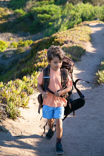 田舎道を歩いて、巨大なバックパックを運ぶかわいい男の子。正面図、全長。子供の頃または冒険旅行の概念