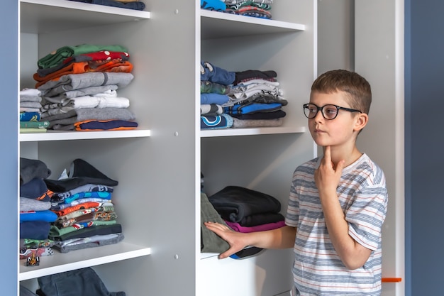 A cute boy with glasses stands near a wardrobe and thinks about what to wear.