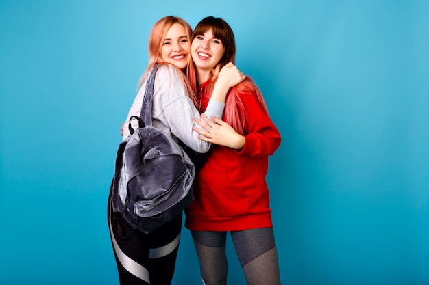 Free photo cute bright portrait of two happy pretty hipster girls wearing sportive clothes for fitness and backpack, smiling and hugs, blue wall.