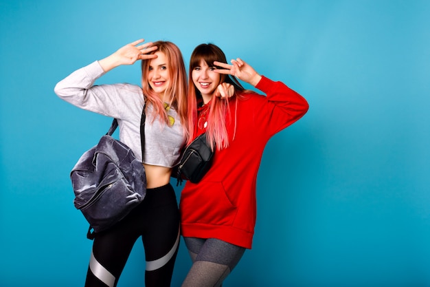 Милый яркий портрет двух счастливых симпатичных хипстерских девушек в спортивной одежде для фитнеса и рюкзака, улыбок и объятий, синяя стена.
