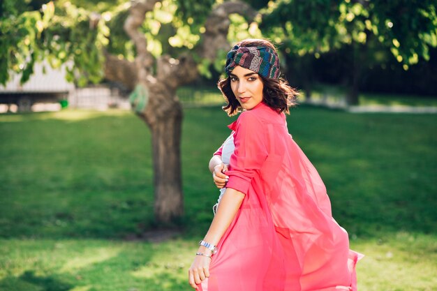 Cute brunette girl in bandana walking in summer park. She wears white clothes, her long pink shirt is flying on wind. View from back.