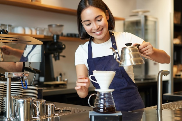 Cute brunette girl barista cafe staff pouring water from kettle and brewing filter coffee behind cou