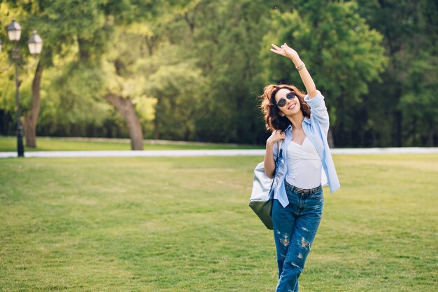 Милая брюнетка девушка с короткими волосами в солнцезащитных очках, позирует в парке. Она носит белую футболку, синюю рубашку и джинсы, сумку. Она держит руку выше и улыбается.
