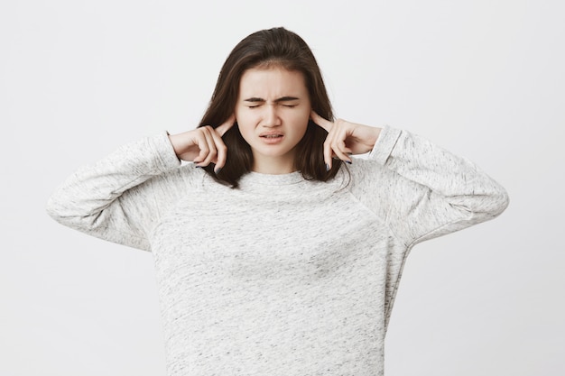 cute brunette woman, expressing stress and pressure, holding fingers in ears not to hear something and closing her eyes