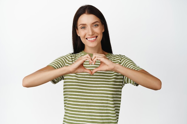 Cute brunette woman shows heart gesture I love you sign smiling tenderly at camera standing over white background in casual outfit