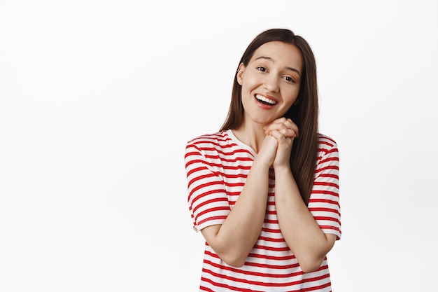 Cute brunette woman sighing, looking at something lovely and touched, clench hands and smiling, gazing and admire smth, standing against white wall