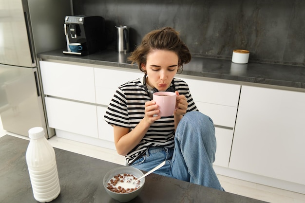 可愛い茶色の女性の学生が朝食を食べ ⁇ 熱いお茶を飲み ⁇ 笑顔で満足しているように見えます ⁇ 