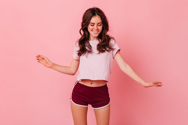 Cute brunette woman in summer outfit of sporty style is having fun dancing on pink wall