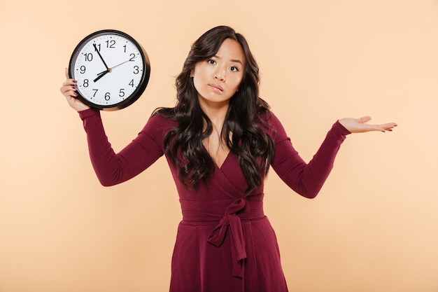 Cute brunette woman with curly long hair holding clock showing nearly 8 being late or missing something throwing up hand over peach background