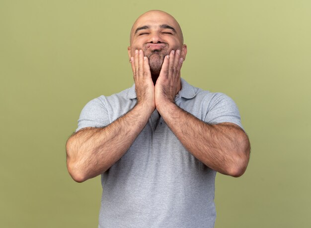 Cute casual middle-aged man keeping hands on cheeks pursing lips with closed eyes isolated on olive green wall