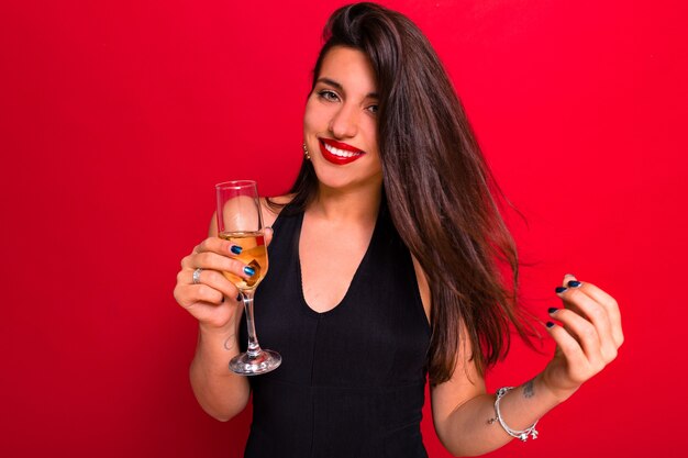 赤い唇と長い黒髪のかわいい白人のエレガントな女性は、ふざけてカメラを見て、シャンパンを飲みます。