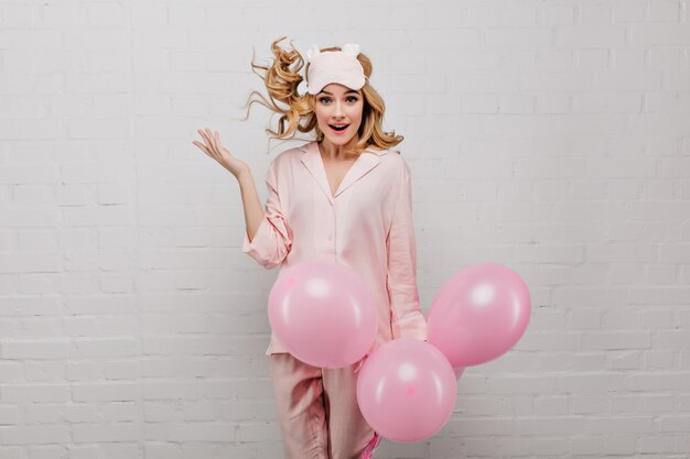 Cute caucasian lady in sleepmask jumping on white wall with party balloons. amazed attractive birthday girl having fun in morning.