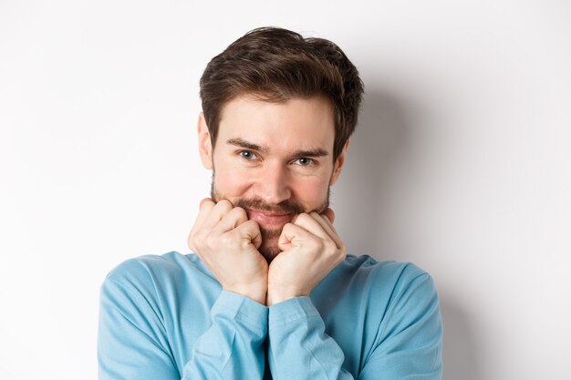 Cute caucasian man looking at something with admiration and desire, touching face with silly smile, standing over white background.