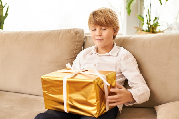 Foto gratuita adolescente caucasico sveglio che si siede sul sofà con il regalo del nuovo anno sulle sue ginocchia. bel ragazzo pronto ad aprire la scatola d'oro con il regalo di natale, con un'espressione facciale anticipata curiosa, sorridente