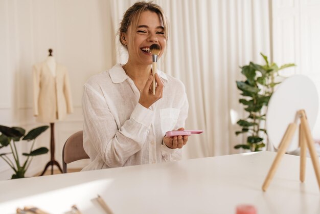 Cute caucasian young blonde girl laughs powders her nose while sitting at table indoors. Cosmetics, makeup finishing, brush.