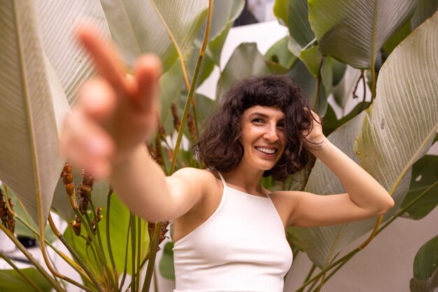 Cute caucasian young brunette girl in summer top reaches out with her hand to camera on background of plants Lifestyle female beauty concept