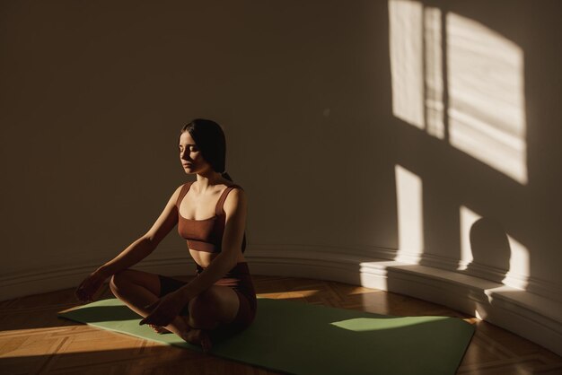 Cute caucasian young woman meditates while sitting on mat near light wall space for text Darkhaired girl wears top and leggings in training Zen concept