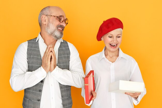 Cute charming middle aged female in stylish headwear opening mouth in excitement while opening box with present from her husband on birthday, bearded senior man rubbing hands with pleasure